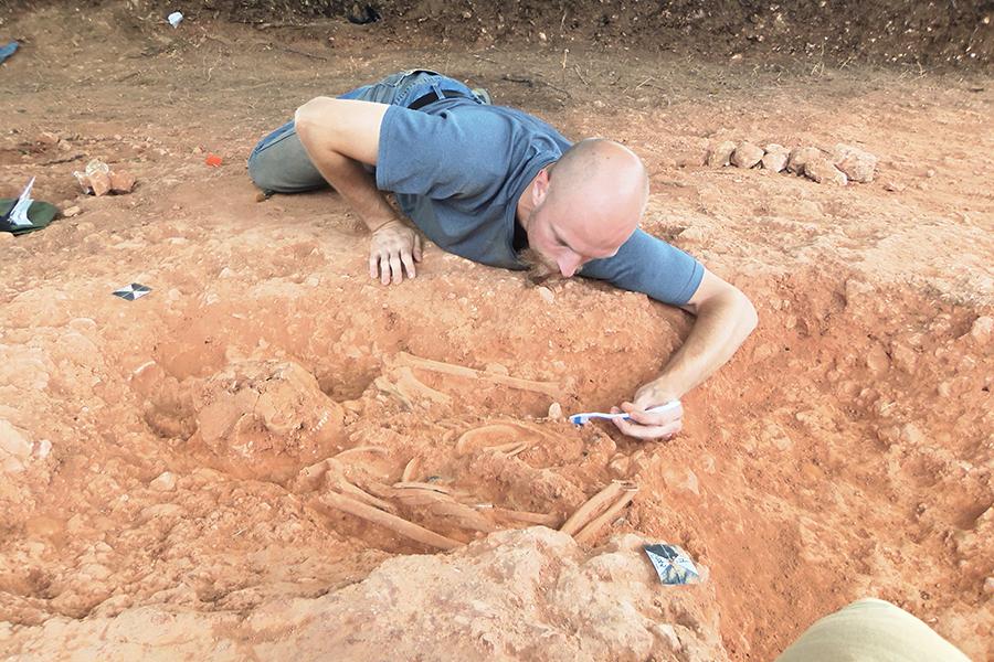 A student lays in the dirt of a dig site and uncovers a skeleton with a toothbrush.